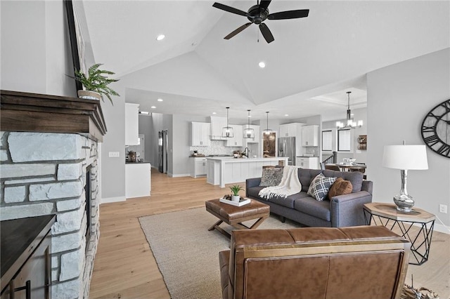 living room with ceiling fan with notable chandelier, vaulted ceiling, a stone fireplace, and light hardwood / wood-style flooring