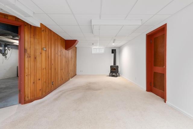 basement featuring light carpet, a paneled ceiling, a wood stove, and wood walls