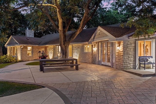 back of property at dusk featuring french doors, a chimney, a shingled roof, a patio area, and stone siding