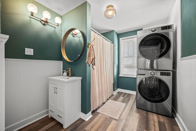 washroom with a sink, a wainscoted wall, stacked washer / drying machine, and wood finished floors