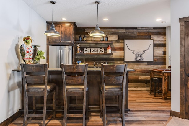 bar with a textured ceiling, wood-type flooring, hanging light fixtures, and stainless steel refrigerator