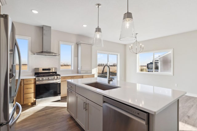 kitchen featuring wall chimney range hood, sink, appliances with stainless steel finishes, a kitchen island with sink, and decorative light fixtures