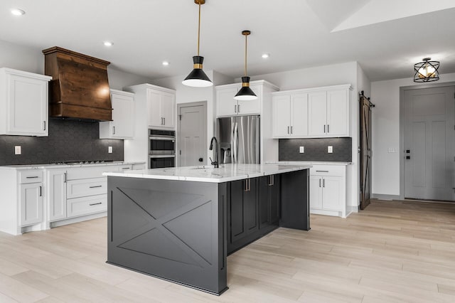 kitchen featuring appliances with stainless steel finishes, a barn door, white cabinets, hanging light fixtures, and an island with sink