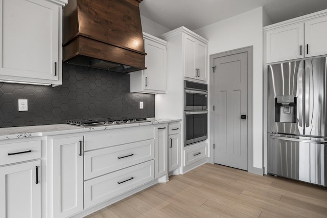 kitchen featuring white cabinets, light stone countertops, light wood-type flooring, appliances with stainless steel finishes, and custom range hood