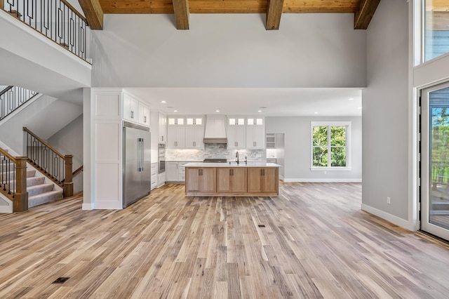 kitchen featuring white cabinetry, beamed ceiling, premium range hood, a large island with sink, and built in refrigerator