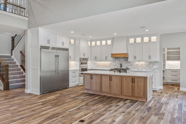 kitchen featuring white cabinets, custom exhaust hood, an island with sink, and appliances with stainless steel finishes