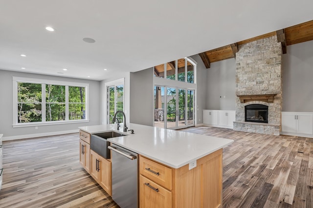 kitchen featuring light brown cabinetry, stainless steel dishwasher, a fireplace, vaulted ceiling with beams, and an island with sink