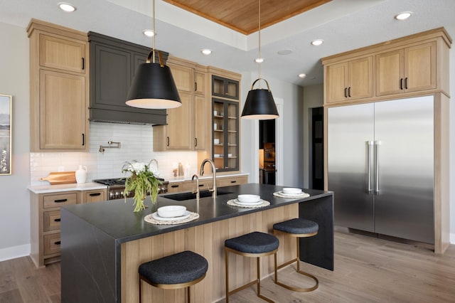 kitchen with sink, built in refrigerator, a kitchen island with sink, tasteful backsplash, and a tray ceiling