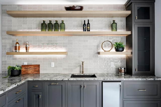 bar featuring stainless steel refrigerator, sink, gray cabinetry, backsplash, and light stone countertops