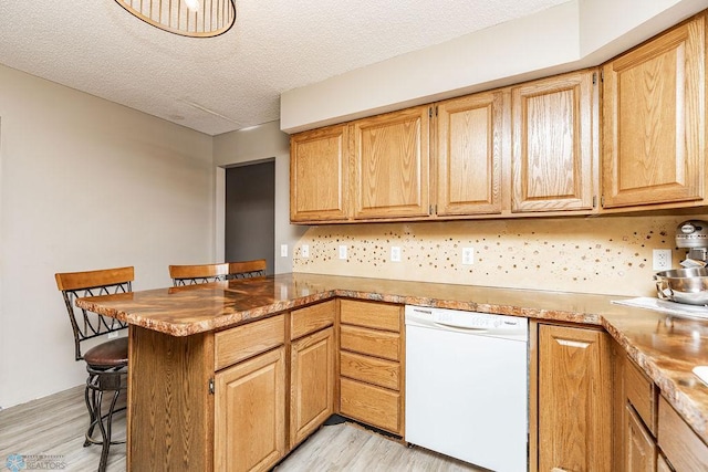 kitchen featuring light wood finished floors, a kitchen breakfast bar, a peninsula, white dishwasher, and a textured ceiling