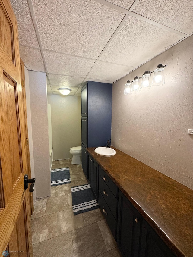 full bathroom featuring stone tile flooring, a drop ceiling, vanity, and toilet