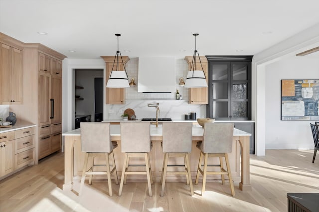 kitchen with pendant lighting, light hardwood / wood-style floors, custom range hood, and an island with sink