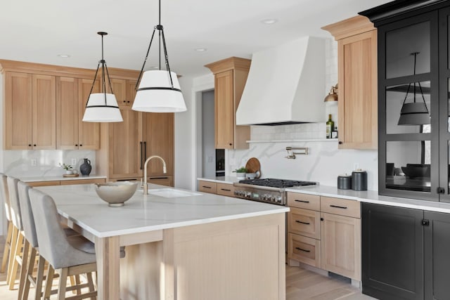 kitchen with hanging light fixtures, premium range hood, an island with sink, decorative backsplash, and light wood-type flooring