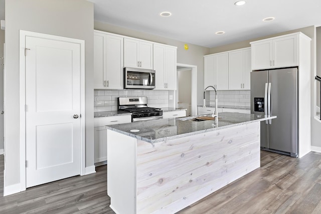 kitchen featuring white cabinets, stainless steel appliances, dark stone counters, and an island with sink