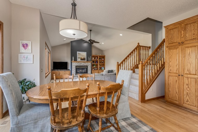 dining space featuring light wood-type flooring, a brick fireplace, a textured ceiling, vaulted ceiling, and ceiling fan