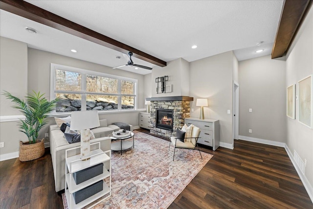 living room with beam ceiling, dark wood-type flooring, a stone fireplace, and ceiling fan