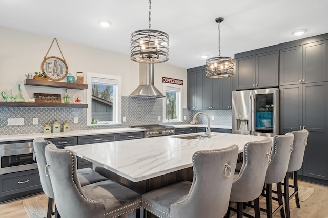 kitchen featuring wall chimney range hood, sink, stainless steel fridge, light stone counters, and an island with sink