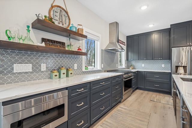 kitchen with decorative backsplash, stainless steel appliances, light stone countertops, light wood-type flooring, and wall chimney exhaust hood