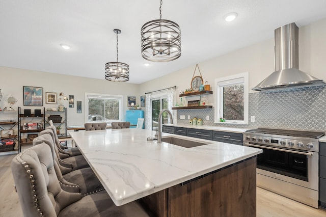 kitchen featuring wall chimney range hood, sink, a kitchen island with sink, stainless steel range, and decorative light fixtures