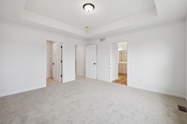 unfurnished bedroom featuring a raised ceiling, a spacious closet, light colored carpet, and ensuite bath