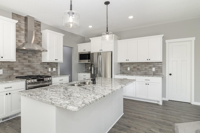 kitchen with wall chimney range hood, an island with sink, appliances with stainless steel finishes, decorative light fixtures, and dark hardwood / wood-style flooring