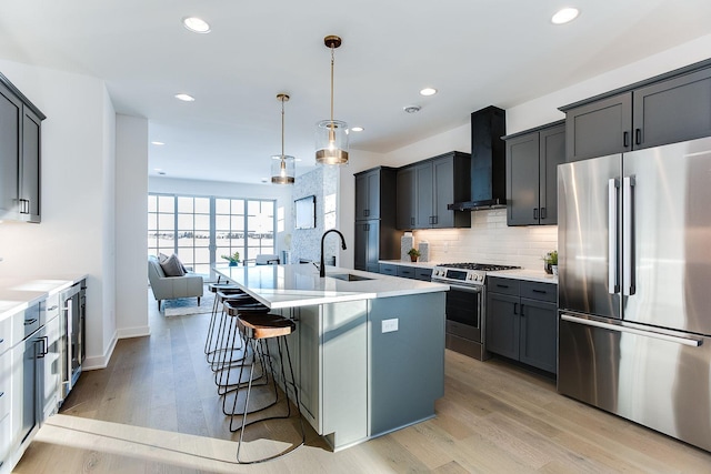 kitchen with appliances with stainless steel finishes, wall chimney exhaust hood, a kitchen island with sink, sink, and pendant lighting