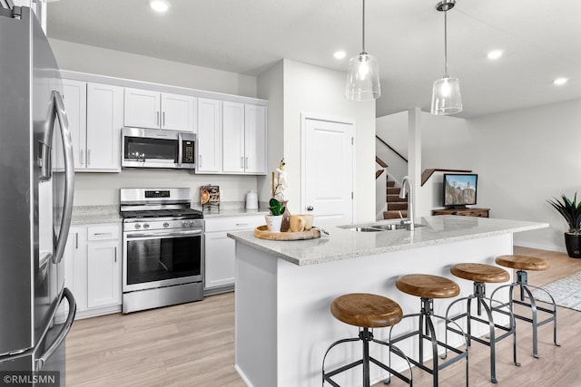 kitchen with hanging light fixtures, stainless steel appliances, an island with sink, white cabinets, and light wood-type flooring