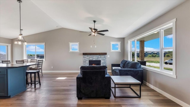 living room featuring ceiling fan with notable chandelier, a stone fireplace, dark wood-type flooring, and vaulted ceiling