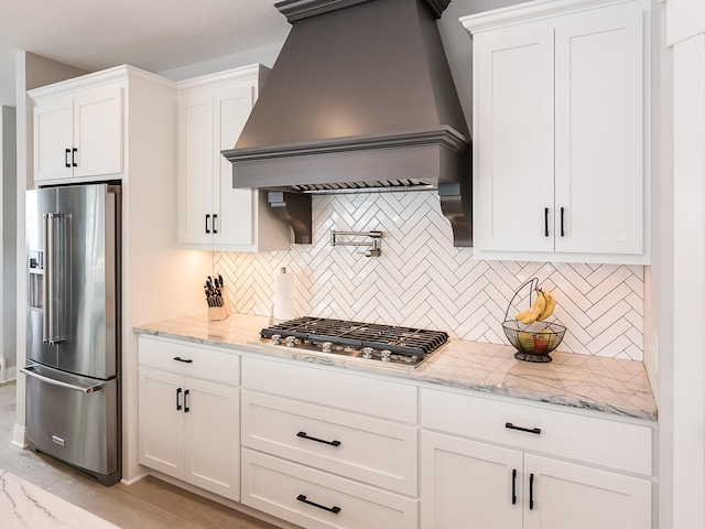 kitchen featuring appliances with stainless steel finishes, backsplash, premium range hood, light stone counters, and white cabinetry