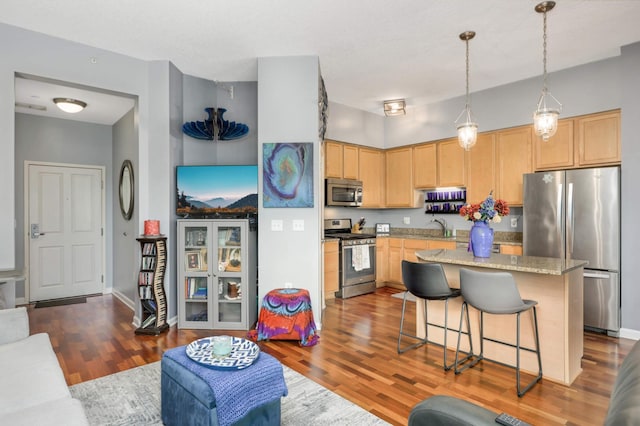 kitchen featuring appliances with stainless steel finishes, light stone counters, light brown cabinets, hardwood / wood-style floors, and hanging light fixtures