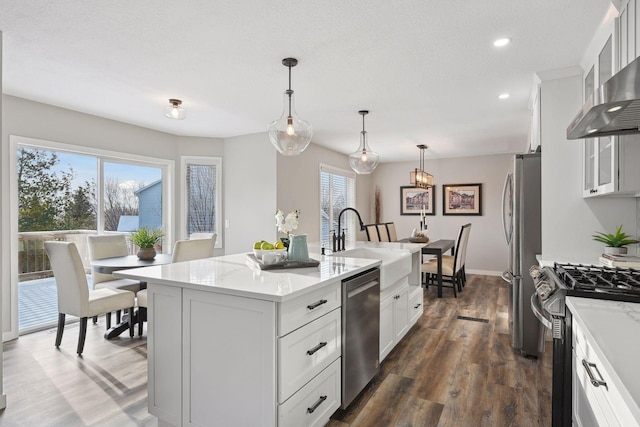 kitchen with wall chimney exhaust hood, stainless steel appliances, dark hardwood / wood-style floors, pendant lighting, and a center island with sink