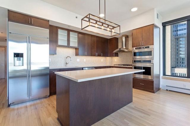 kitchen featuring a center island, wall chimney range hood, light wood-type flooring, appliances with stainless steel finishes, and decorative light fixtures