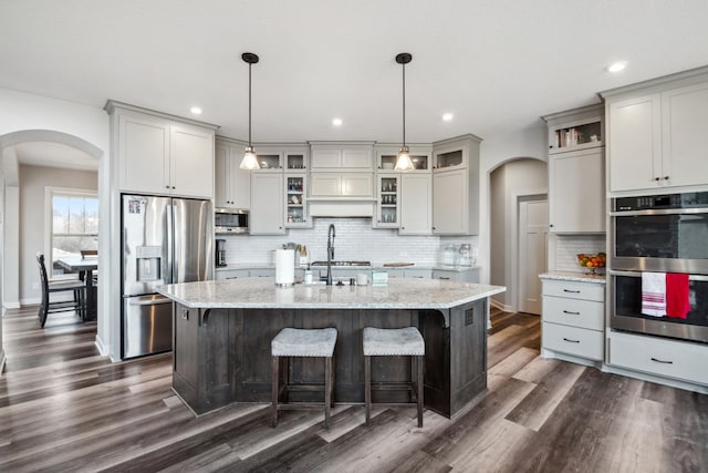 kitchen featuring an island with sink, light stone countertops, arched walkways, and stainless steel appliances