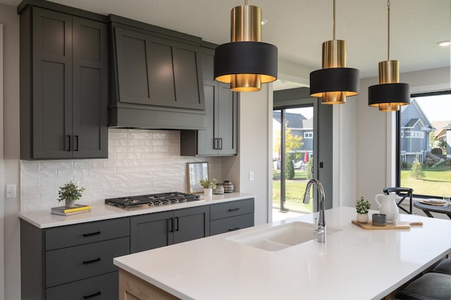 kitchen with pendant lighting, stainless steel gas stovetop, sink, gray cabinets, and an island with sink