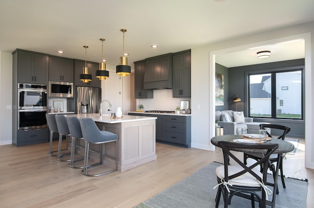 kitchen with premium range hood, decorative light fixtures, a center island with sink, appliances with stainless steel finishes, and light wood-type flooring