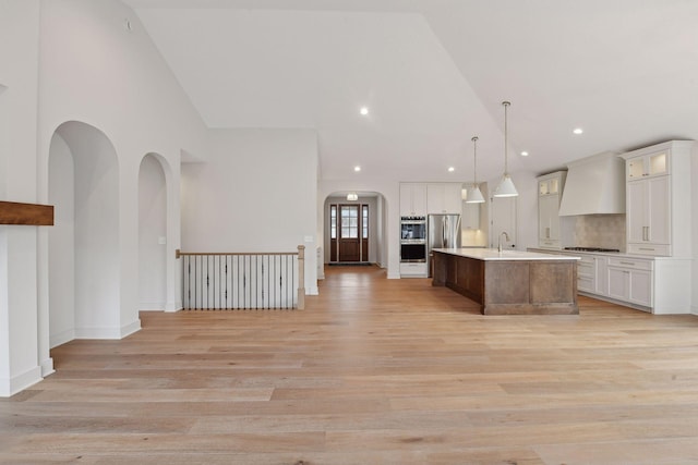 kitchen with custom range hood, stainless steel appliances, a kitchen island with sink, light hardwood / wood-style flooring, and hanging light fixtures