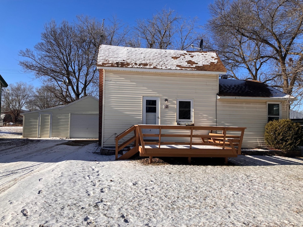 snow covered house with a garage, a deck, and an outbuilding