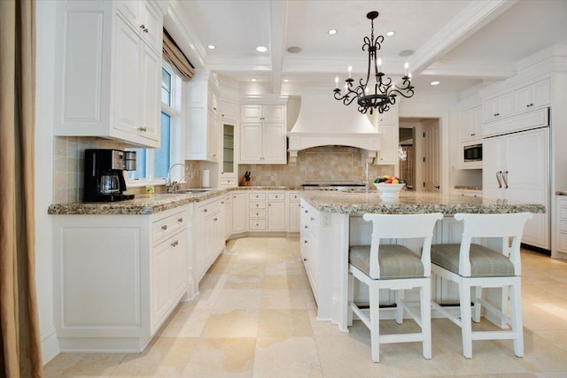 kitchen with pendant lighting, premium range hood, a kitchen island with sink, white cabinetry, and light stone counters