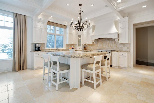 kitchen with premium range hood, white cabinets, high end range, light stone counters, and a center island with sink