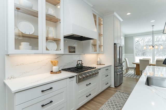 kitchen with a wainscoted wall, stainless steel appliances, white cabinets, custom exhaust hood, and decorative light fixtures