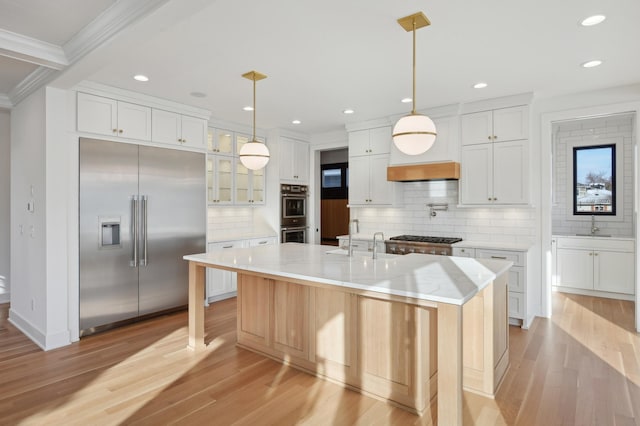 kitchen with appliances with stainless steel finishes, ornamental molding, a kitchen island with sink, light hardwood / wood-style flooring, and white cabinetry