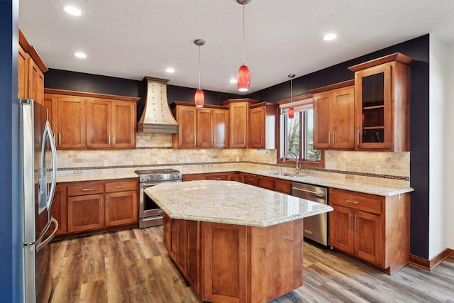 kitchen with custom exhaust hood, hanging light fixtures, appliances with stainless steel finishes, a kitchen island, and wood-type flooring