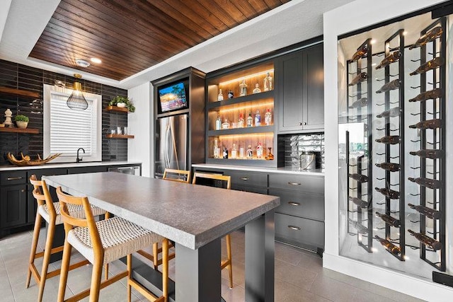 bar featuring tile patterned floors, backsplash, stainless steel refrigerator, and wooden ceiling