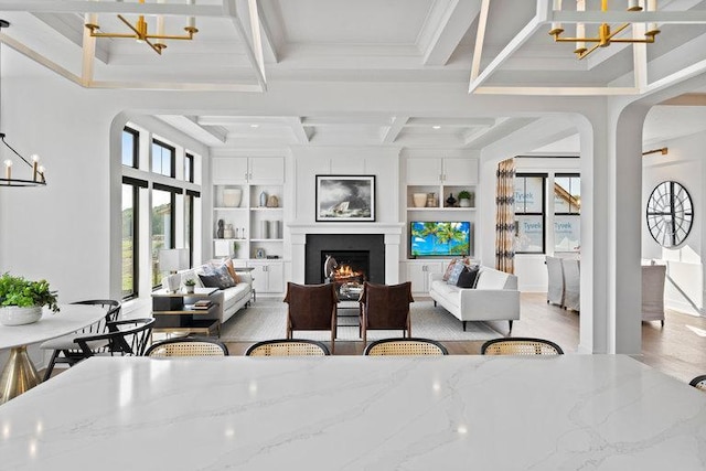 living area featuring built in shelves, arched walkways, an inviting chandelier, a warm lit fireplace, and coffered ceiling