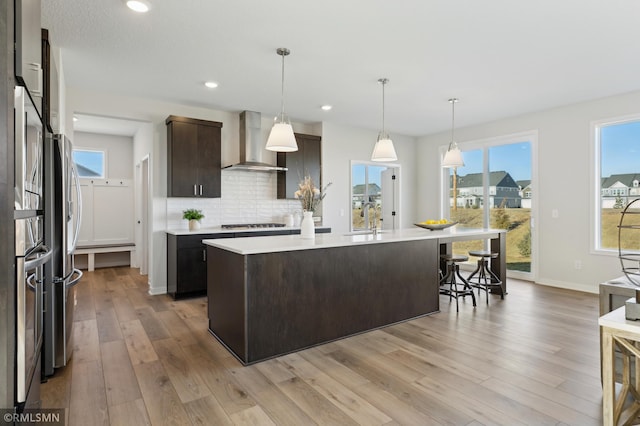 kitchen featuring tasteful backsplash, wall chimney range hood, dark brown cabinetry, hanging light fixtures, and an island with sink