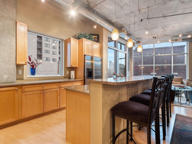 kitchen featuring stainless steel refrigerator with ice dispenser, light wood-type flooring, track lighting, light stone counters, and a center island with sink