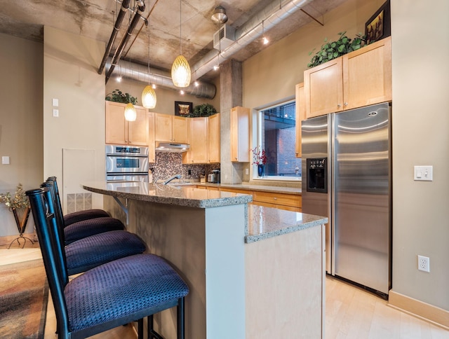 kitchen with light brown cabinets, stainless steel appliances, hanging light fixtures, and an island with sink