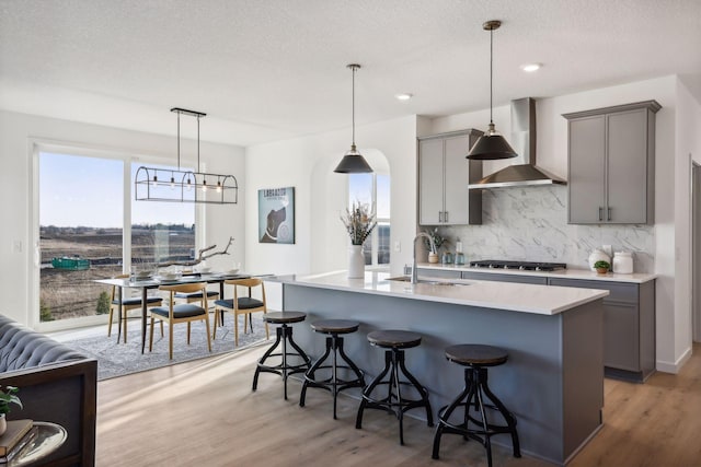 kitchen with gray cabinetry, sink, wall chimney range hood, hardwood / wood-style floors, and a kitchen island with sink