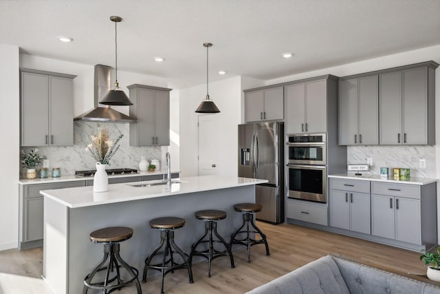 kitchen with gray cabinets, a kitchen island with sink, stainless steel appliances, and wall chimney range hood