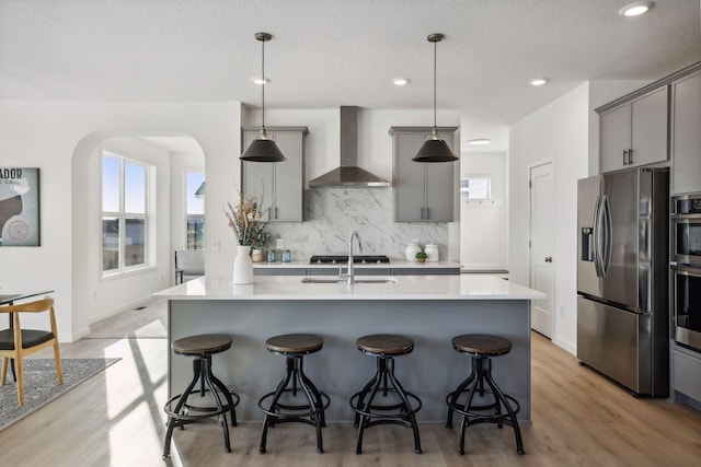 kitchen with light wood-type flooring, wall chimney exhaust hood, stainless steel appliances, gray cabinets, and hanging light fixtures